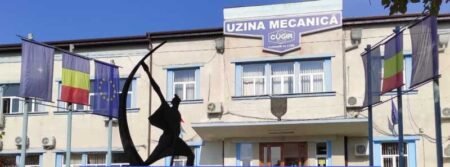 Фасад завода Uzina Mecanică в Кужире, Румыния, с флагами и памятником на переднем плане