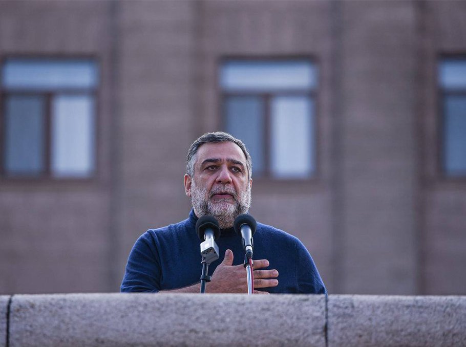 Мужчина с седой бородой в тёмном свитере выступает за каменной трибуной с микрофонами на фоне здания