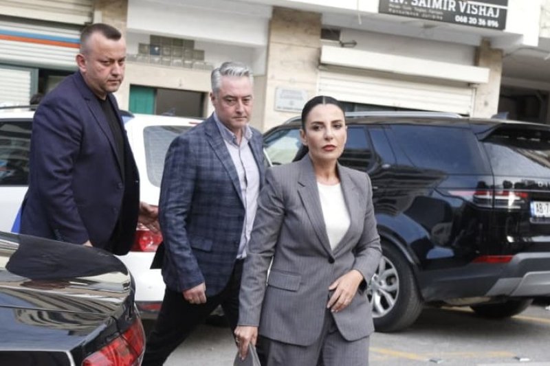 Белинда Баллуку в сером деловом костюме выходит из автомобиля в сопровождении двух мужчин в Тиране