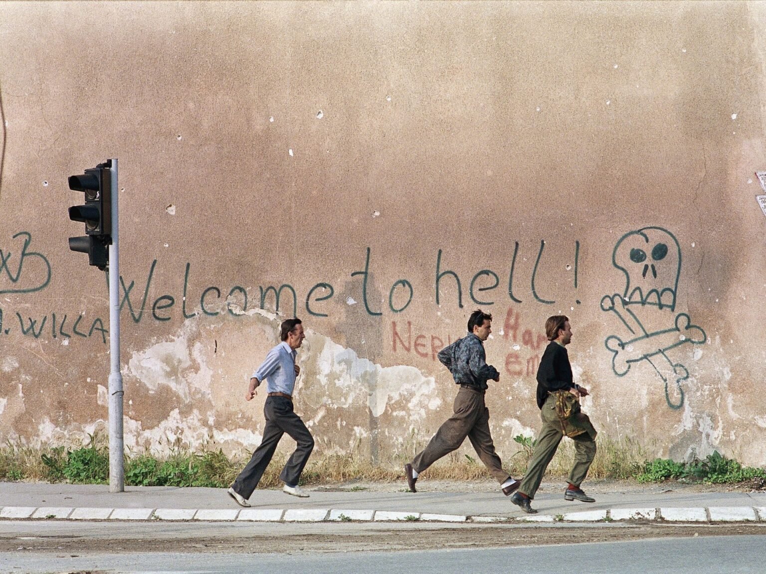 Документы против Вучича о «Сараевском сафари» объявлены фальшивыми — но расследование продолжается Три человека бегут по улице Сараево мимо стены с граффити 'Welcome to hell' и черепом во время осады города в 1993 году