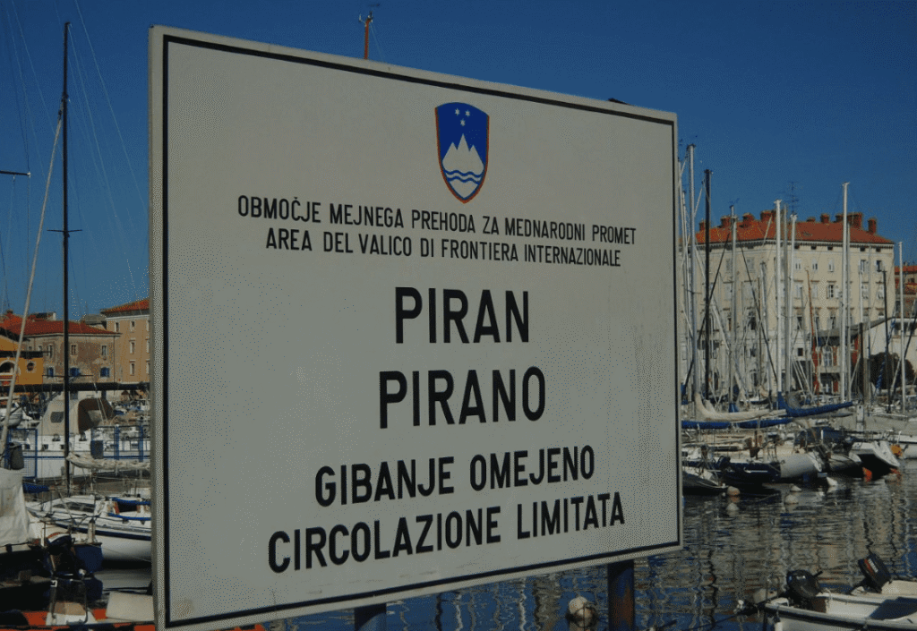 Двуязычная вывеска ПИРАН/PIRANO в гавани Пирана с гербом Словении, демонстрирующая одинаковый размер шрифта для словенского и итальянского языков