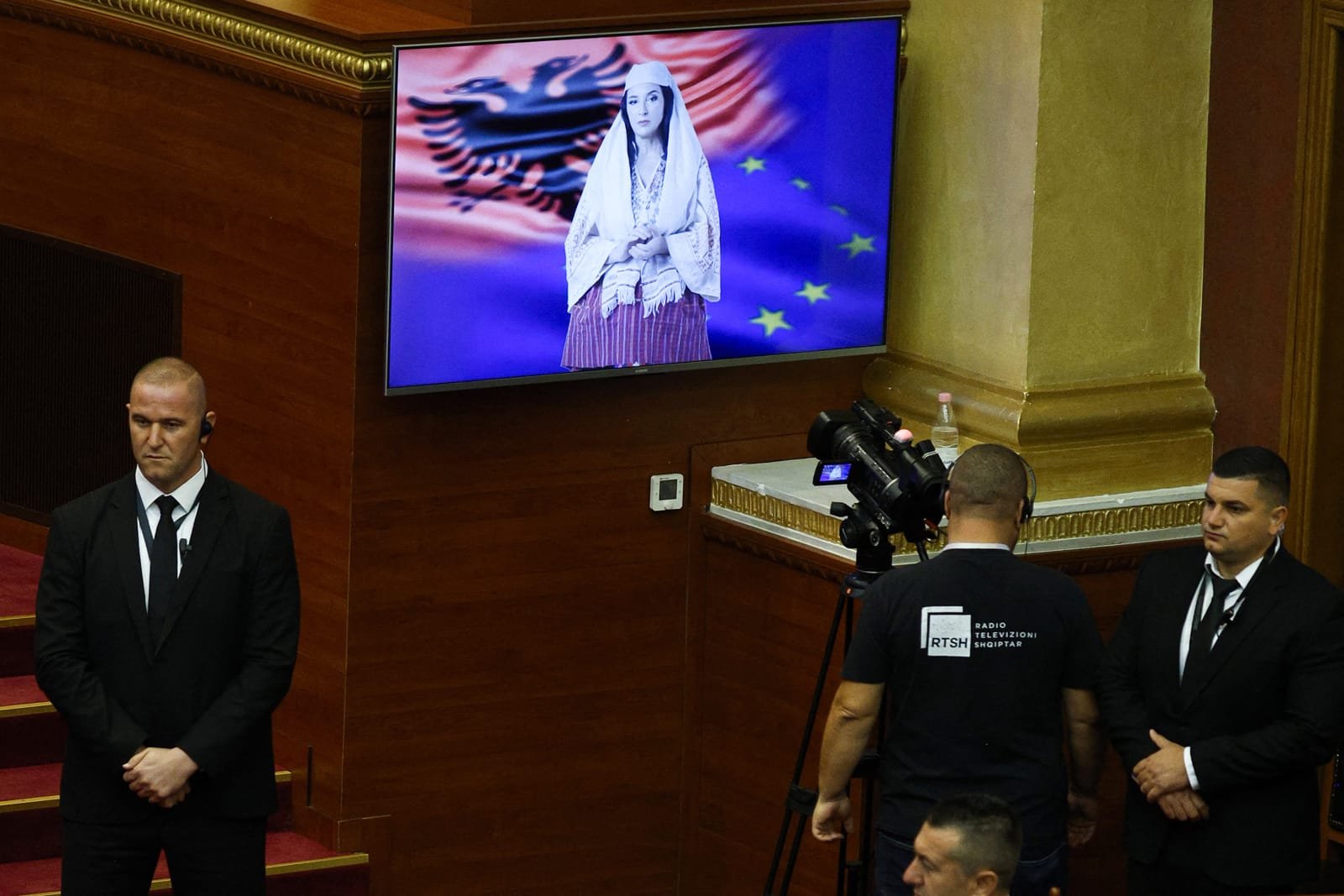 Виртуальный ИИ-министр Диелла на экране в зале парламента Албании на фоне флагов Албании и ЕС, рядом — охрана и оператор RTSH. Фото: Lowy Institute / AFP