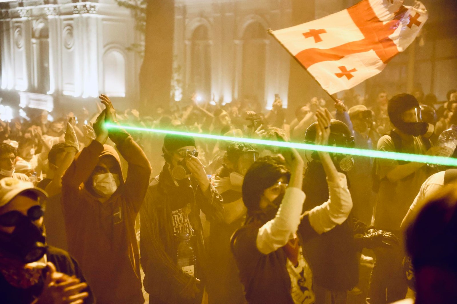 Nighttime protest scene with protesters in gas masks, Georgian flag waving, green laser beam cutting through tear gas haze near parliament