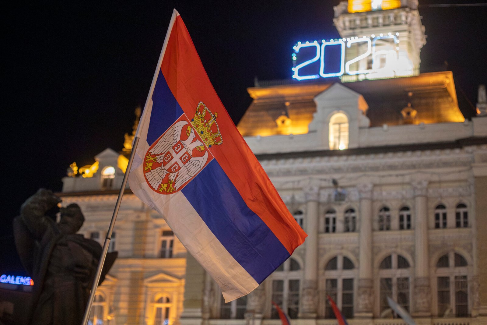 Serbian flag waving at night in front of an illuminated historic building with '2026' sign