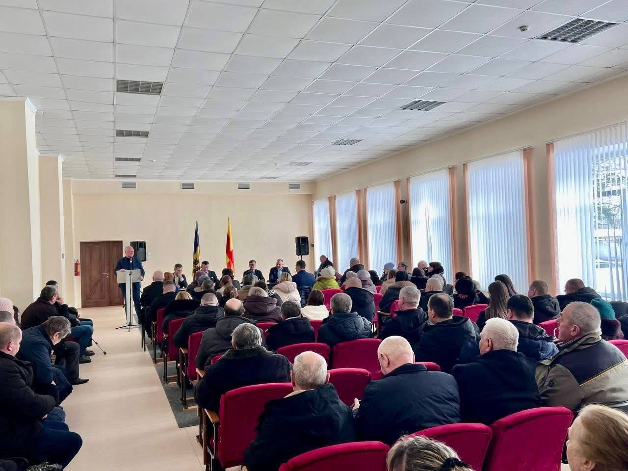 Regional party conference hall with seated audience in red chairs, speaker at podium, flags visible