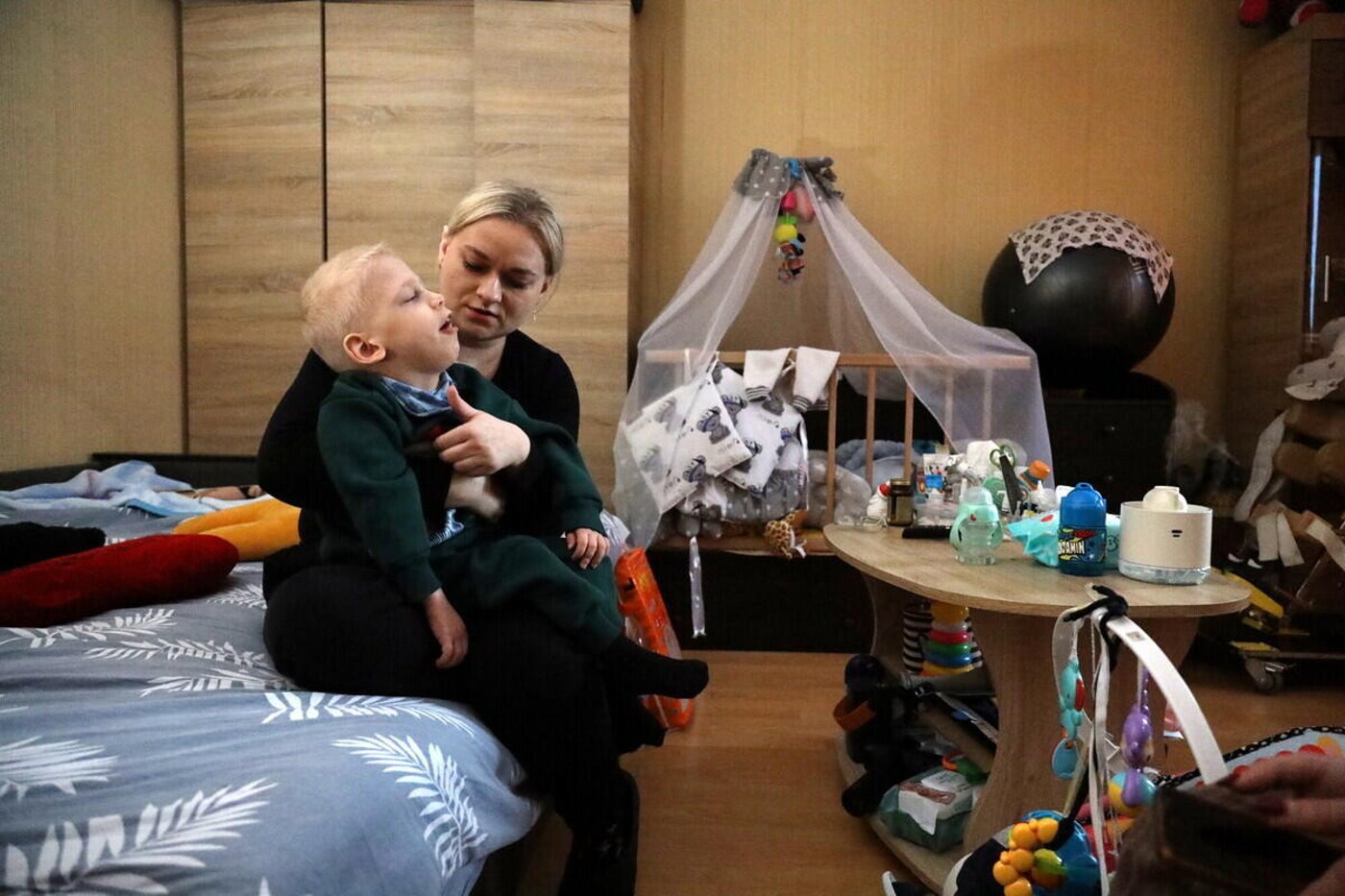 Ukrainian refugee mother holding young child in temporary accommodation room with baby crib and toys
