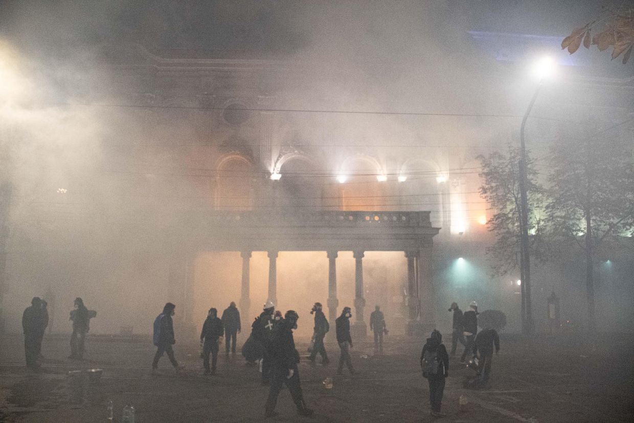 Tear gas engulfs area near Georgian parliament building at night, silhouettes of protesters visible against illuminated columns