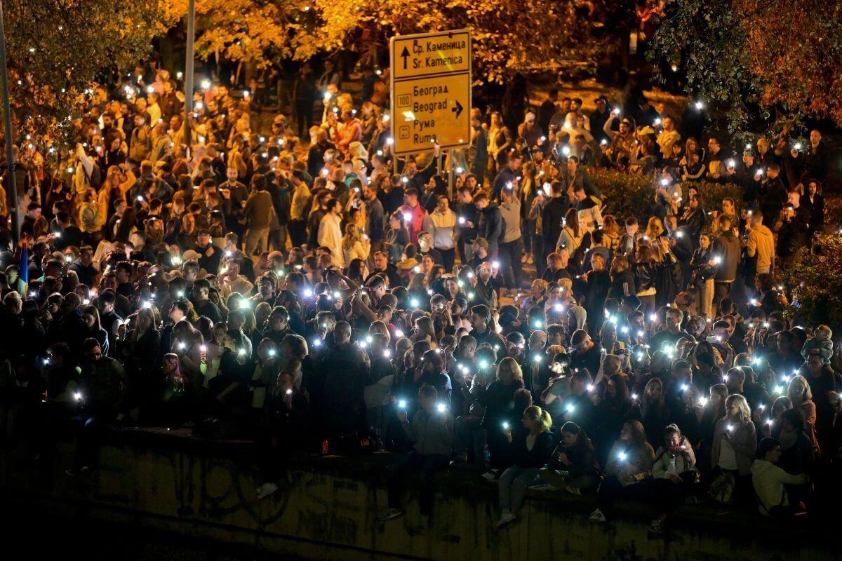 Night protest in Serbia with crowd holding phone flashlights, road signs show Belgrade and Sremska Kamenica