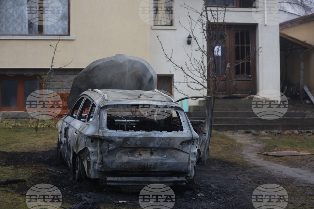 Дневной вид сгоревшего автомобиля перед домом, видны повреждения транспортного средства и входа в здание
