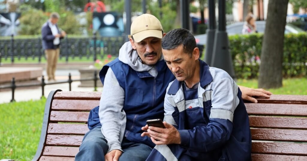 Россия обязывает детей мигрантов уехать в 18 лет Два трудовых мигранта в рабочей одежде сидят на скамейке в парке и смотрят в смартфон
