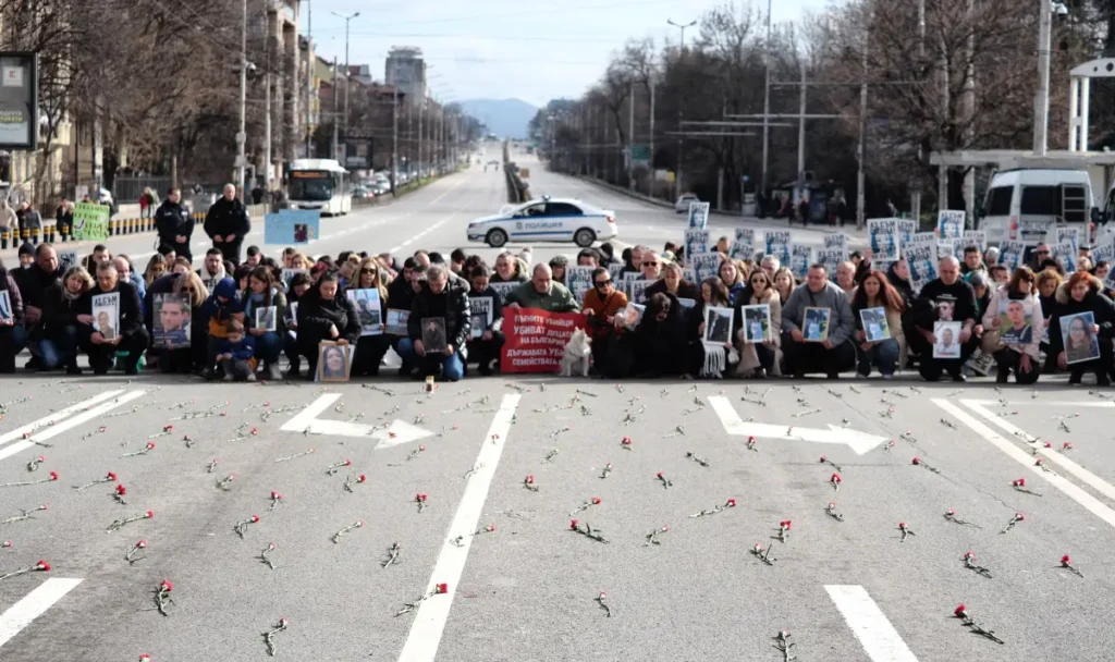 Широкий кадр Орлова моста, перекрытого протестующими, которые стоят на коленях с фотографиями жертв ДТП. 456 красных гвоздик разбросаны по дороге. За живым барьером видна полицейская машина.