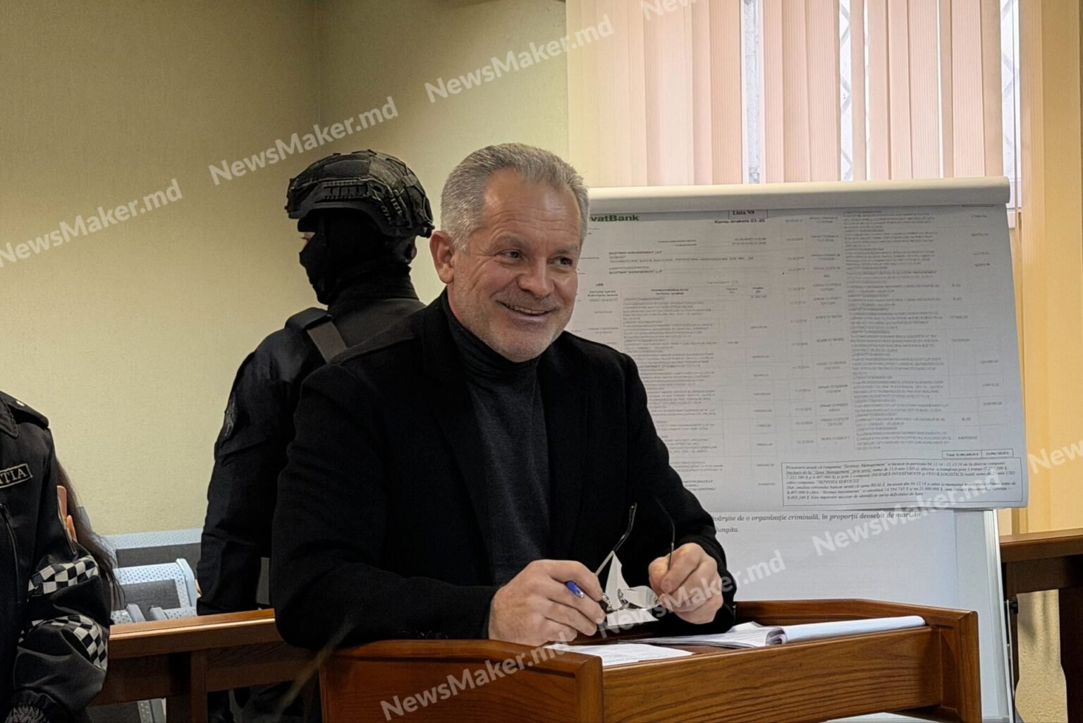 Владимир Плахотнюк улыбается на судебном заседании, за ним диаграммы и вооруженный охранник