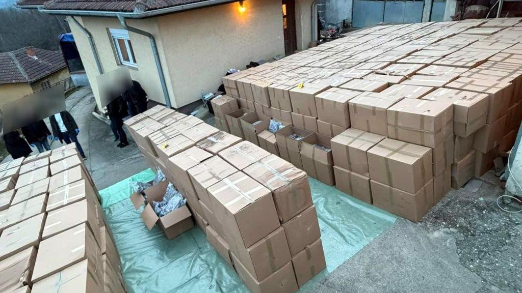 Широкоугольный снимок места обыска: сотни картонных коробок сложены во дворе рядом со зданием, на месте присутствуют сотрудники правоохранительных органов