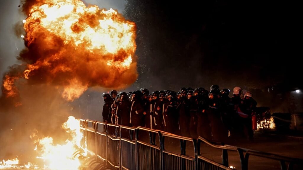 Бойцы спецназа стоят в строю за металлическими ограждениями, пока рядом вспыхивает огромный огненный шар от коктейля Молотова во время ночных столкновений в Тиране