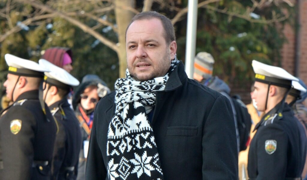 Борислав Сандов на открытом воздухе в черном пальто и шарфе, на фоне военнослужащих