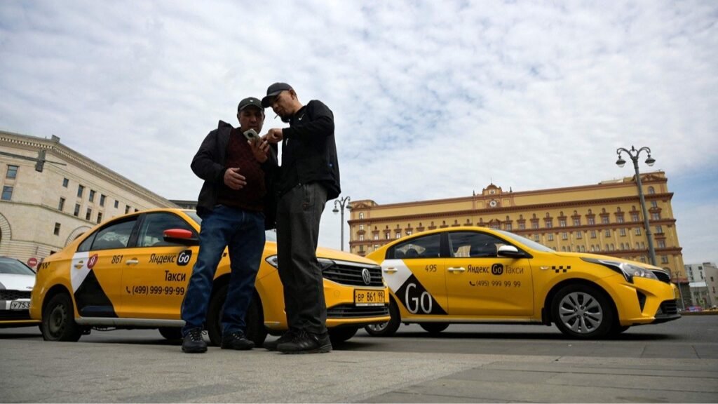 Два водителя такси Яндекс Go стоят у своих желтых машин, проверяют телефон, видна брендинг Яндекса