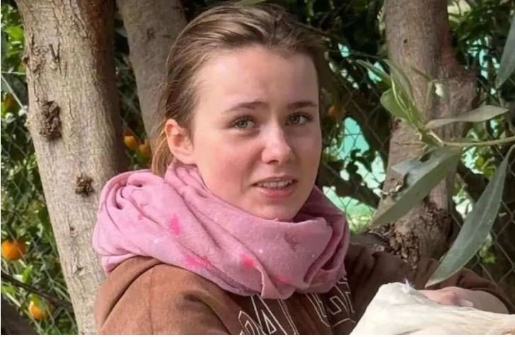 Другое фото Лоры на улице в розовом шарфе и коричневой куртке рядом с деревьями