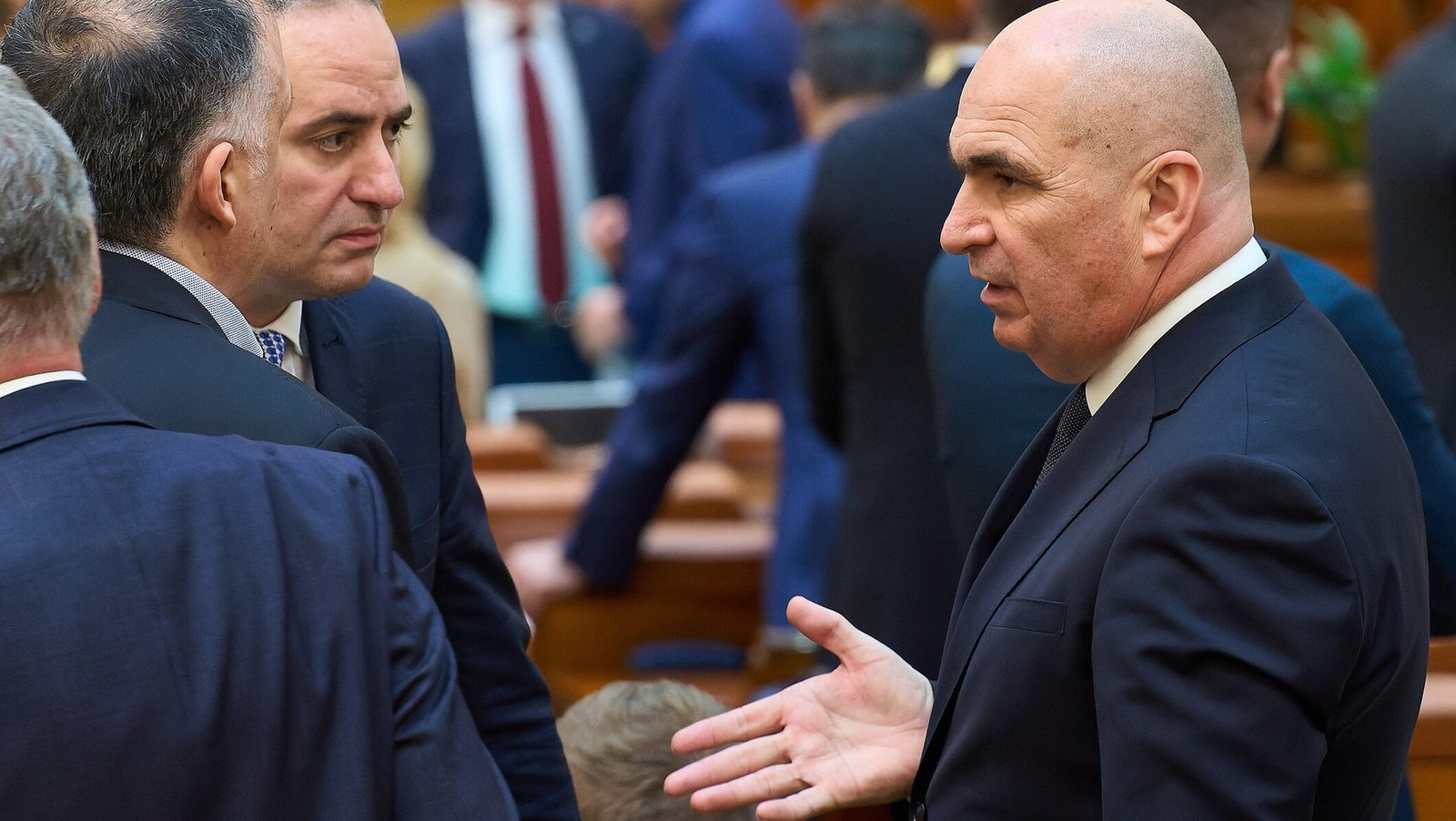 Prime Minister Ilie Bolojan in close conversation with a colleague in the parliament chamber, gesturing with his hand