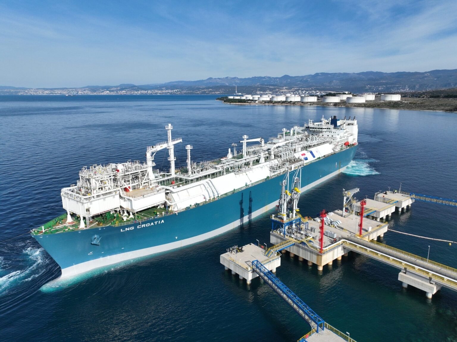 Аэросъёмка СПГ-терминала «LNG Хорватия» на острове Крк: плавучая регазификационная установка у причала, нефтехранилища на полуострове, Адриатическое море и хорватские горы на фоне