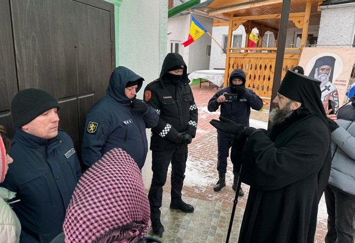 Православный священник в черном облачении разговаривает с молдавскими полицейскими возле церковной территории, виден молдавский флаг