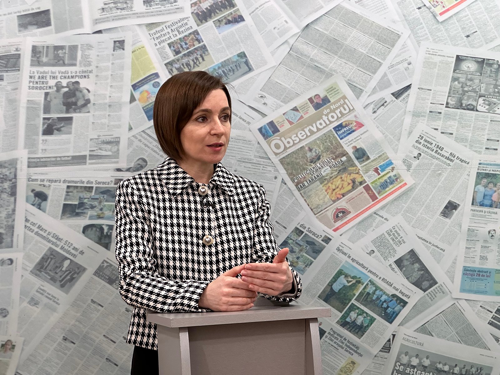 Maia Sandu speaking at podium with wall of newspaper clippings behind her, houndstooth blazer