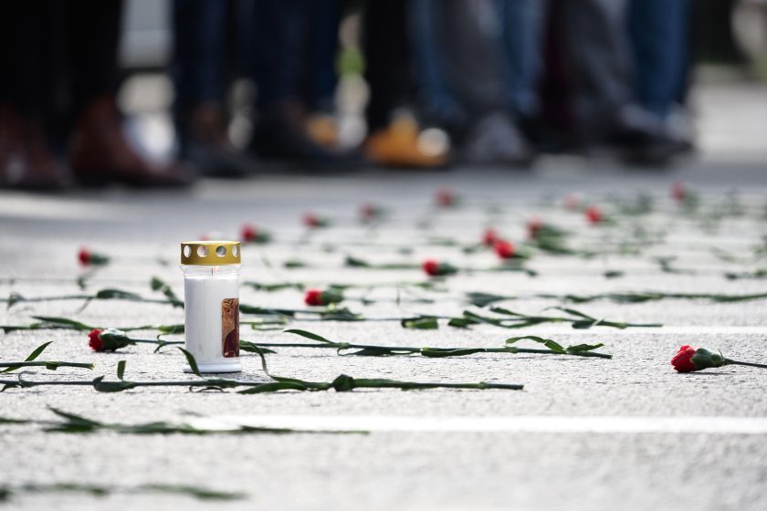 Художественный крупный план: белая поминальная свеча среди разбросанных красных гвоздик на асфальте Орлова моста. На заднем плане — размытые фигуры протестующих.