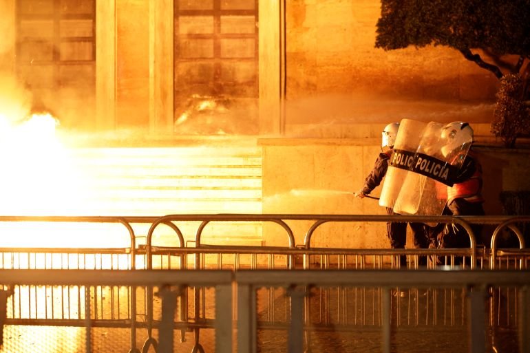 Албанские полицейские с щитами с надписью 'POLICIA' тушат огнетушителем у входа в правительственное здание, пока рядом горит пламя от коктейля Молотова