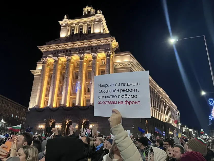 Протестующий с плакатом перед освещённым зданием болгарского парламента ночью