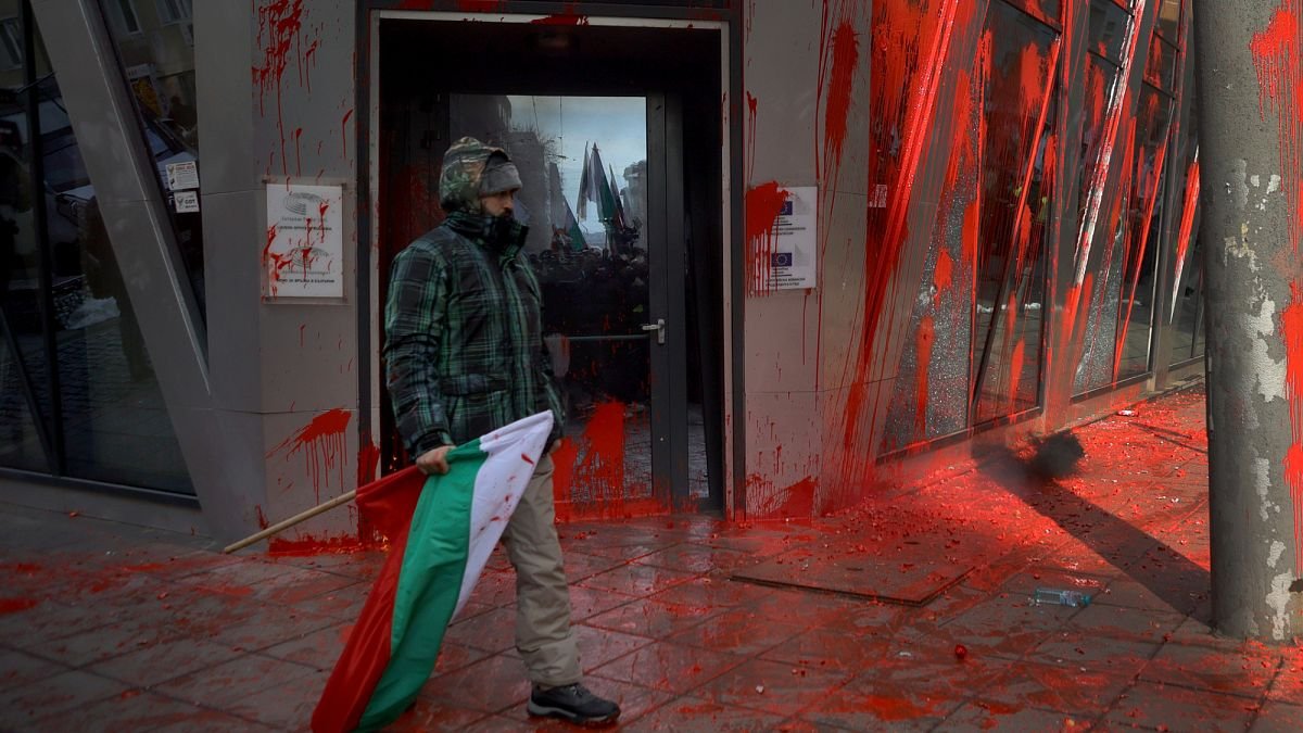 Мужчина с болгарским флагом перед зданием, облитым красной краской, во время политического протеста