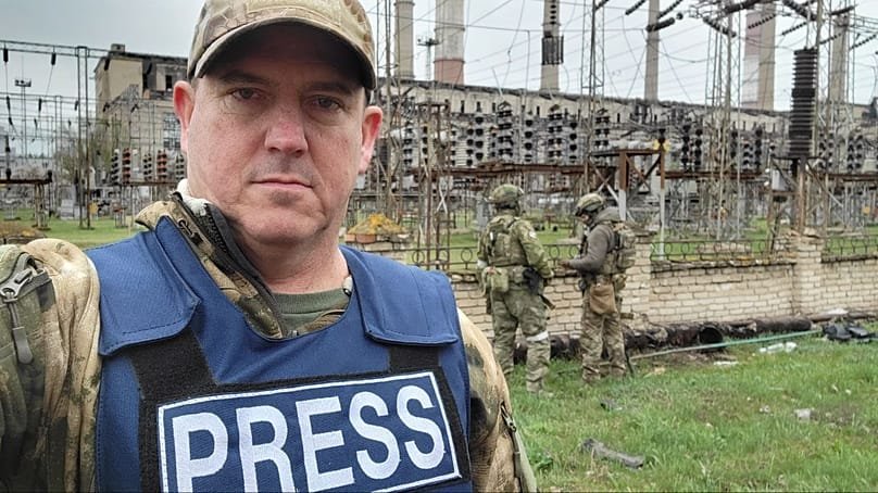 Джон Марк Дуган в бронежилете с надписью PRESS в зоне конфликта — попал под санкции ЕС за распространение российской пропаганды