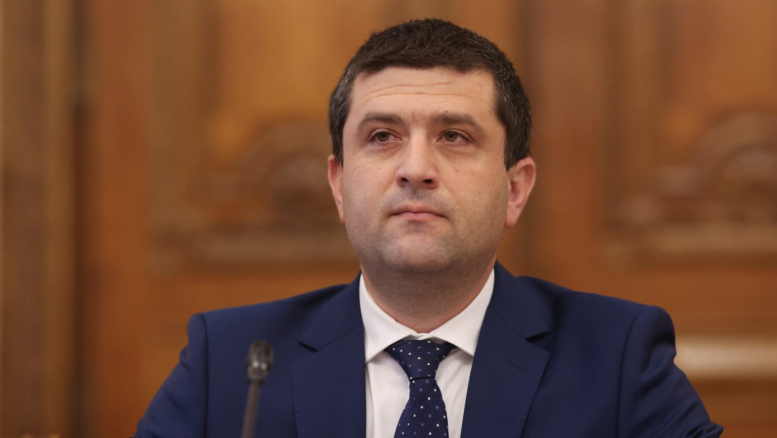 Minister Radu Miruță in formal portrait setting, seated in parliament chamber in a blue suit