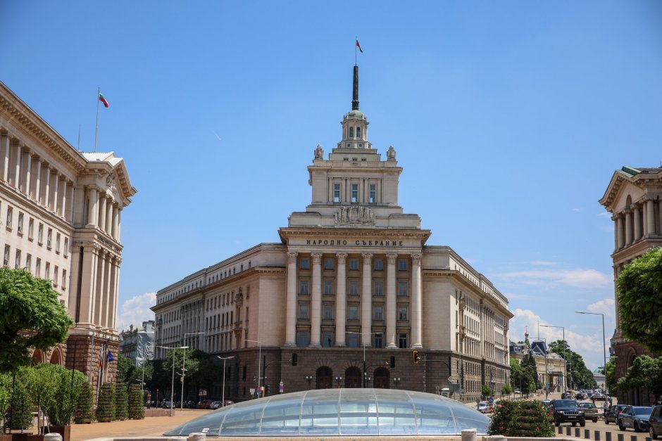 Здание Народного собрания Болгарии в Софии — классическая архитектура с колоннами, флаги Болгарии и ЕС, стеклянный купол на переднем плане.