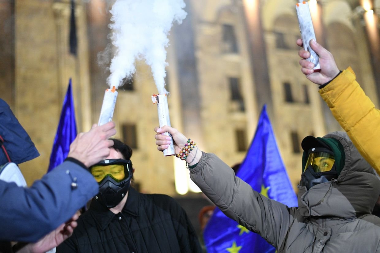 Протестующие в противогазах и очках держат дымовые шашки, на фоне флаги ЕС ночью