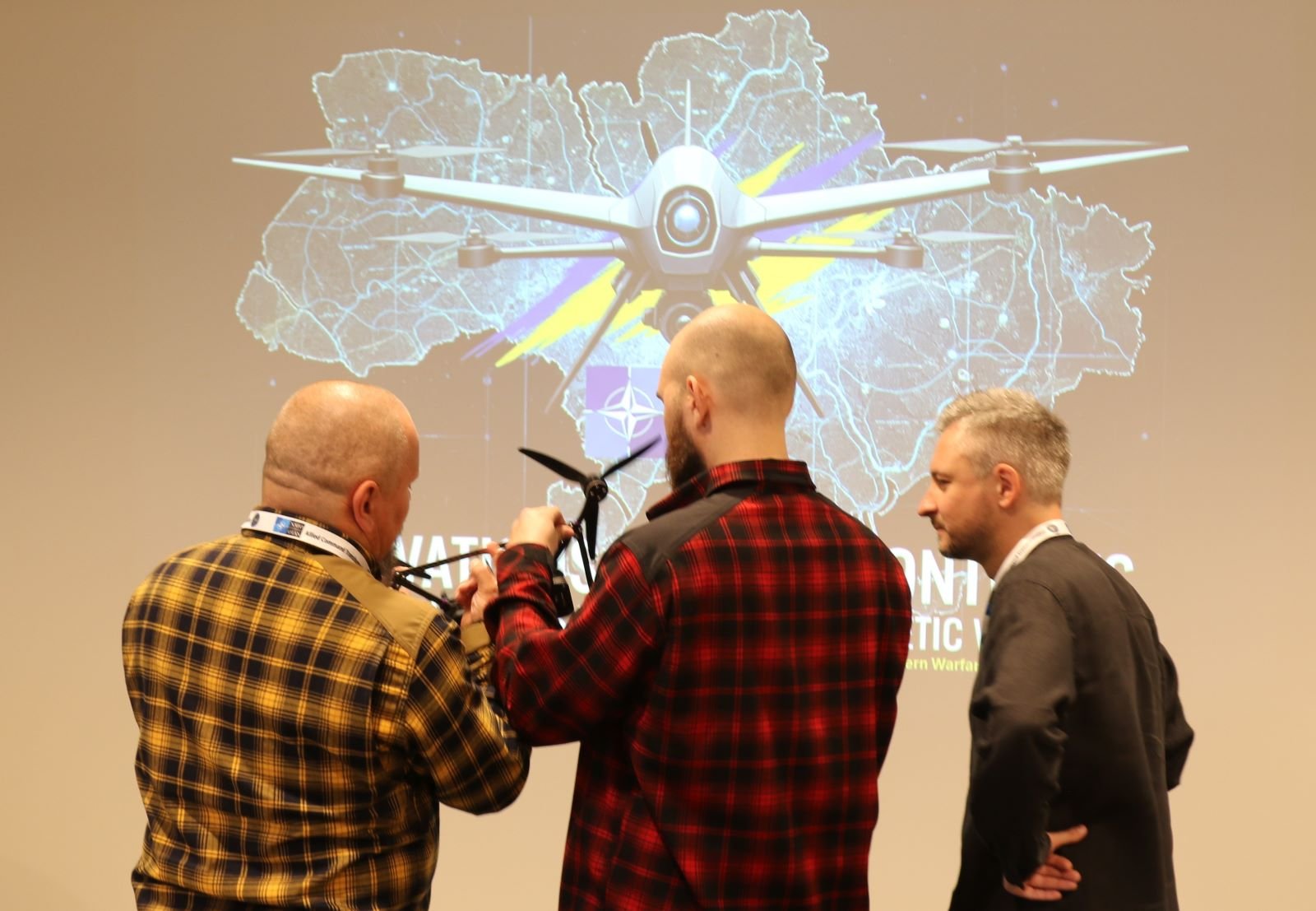 Участники изучают дрон на мероприятии NATO Innovation Challenge на фоне презентации о дроновой войне