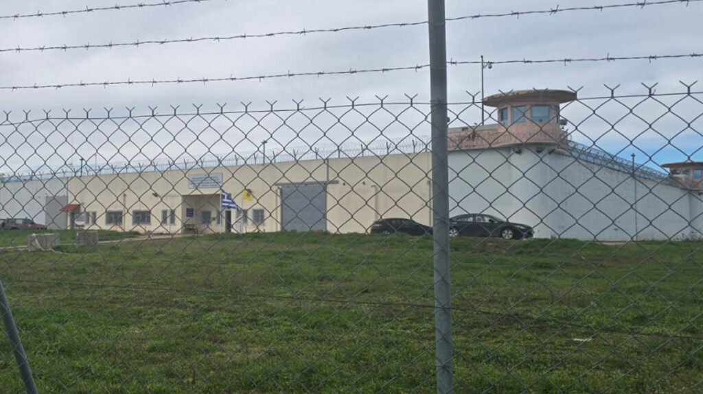 Внешний вид тюрьмы Домокос в Греции: сторожевая вышка, забор с колючей проволокой, тюремные здания и греческий флаг