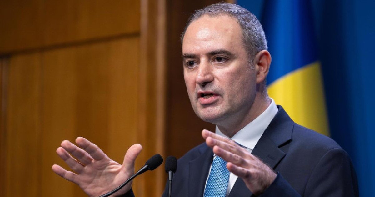 Министр финансов Марчел Болош выступает на пресс-конференции с микрофоном, на заднем плане флаг Румынии