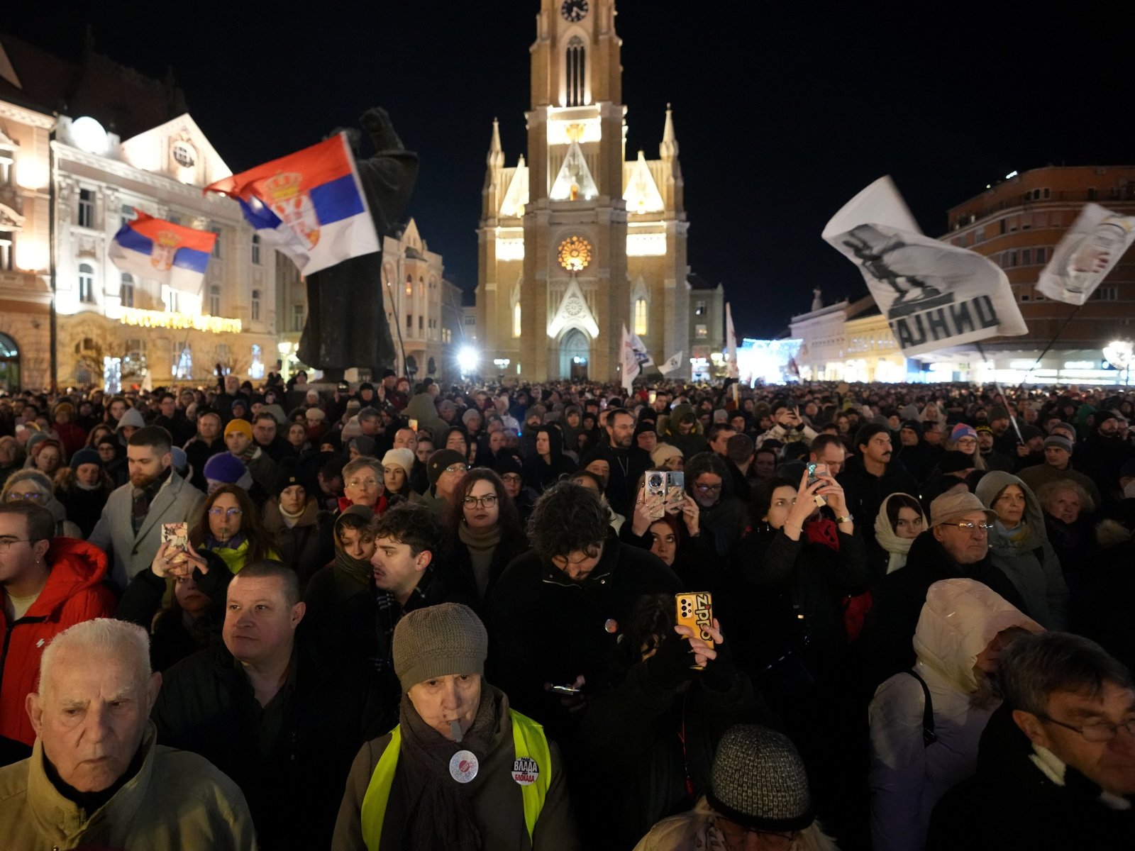 Массовый ночной протест в сербском городе с толпами, размахивающими сербскими флагами перед освещённым собором