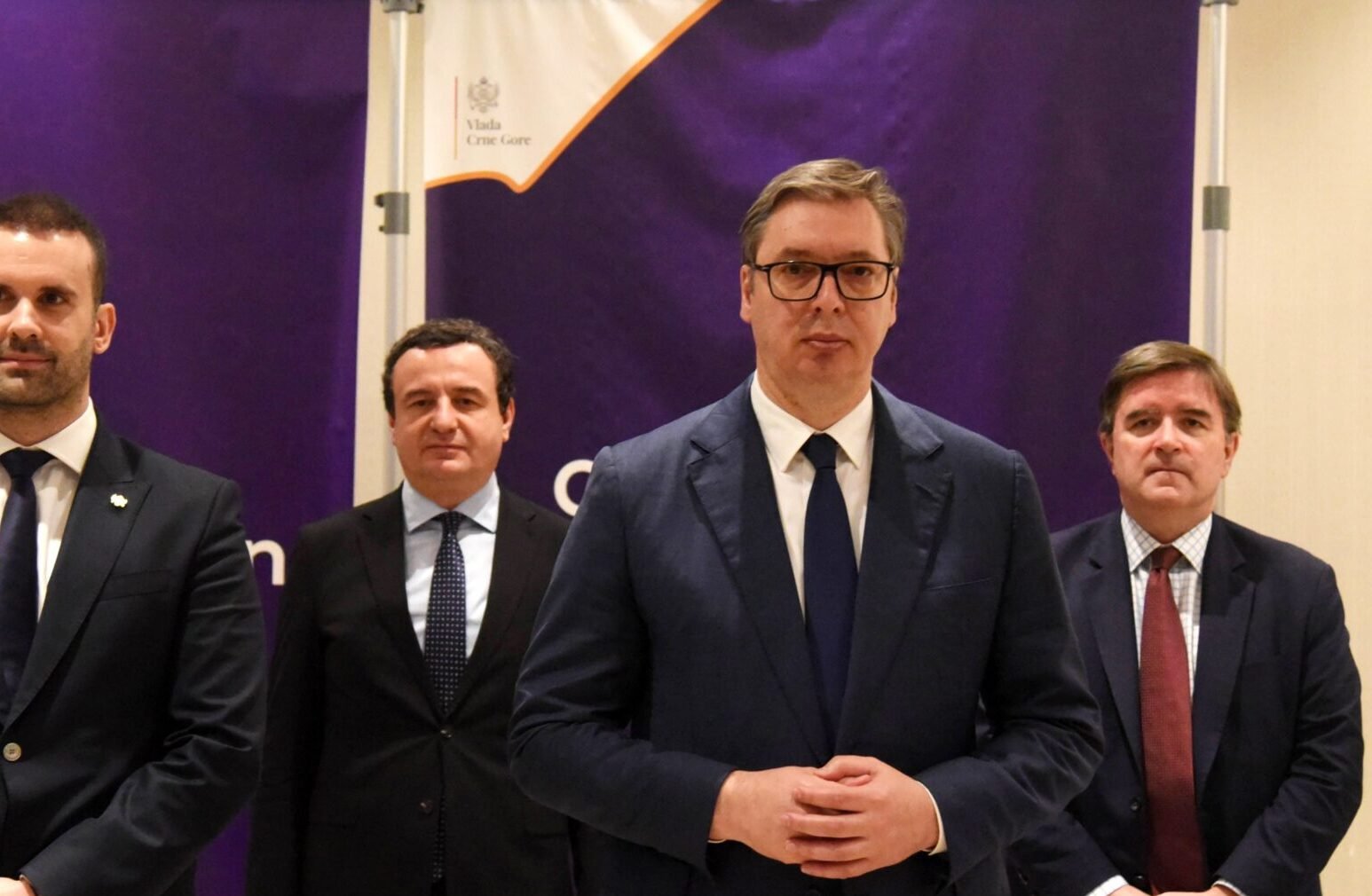 Четверо политиков в костюмах стоят вместе на официальной встрече, за ними виден фиолетовый правительственный баннер