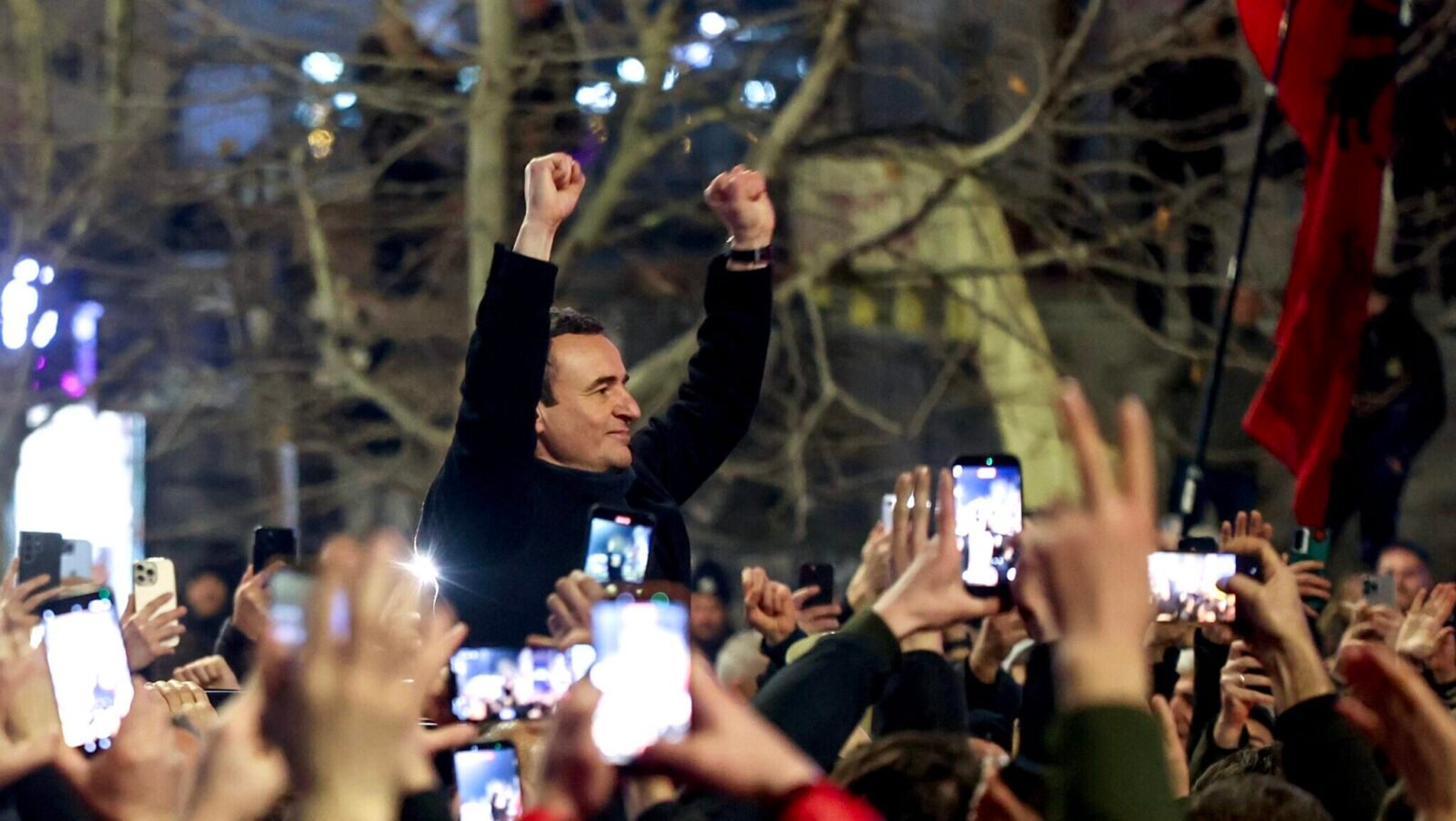 Политический лидер триумфально поднимает кулаки на ночном митинге, пока толпа держит смартфоны, на фоне виден албанский флаг
