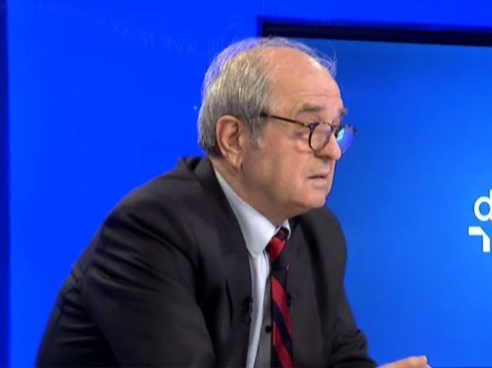 Пожилой мужчина с седыми волосами и в очках в тёмном костюме и полосатом галстуке во время телеинтервью на фоне синей студии
