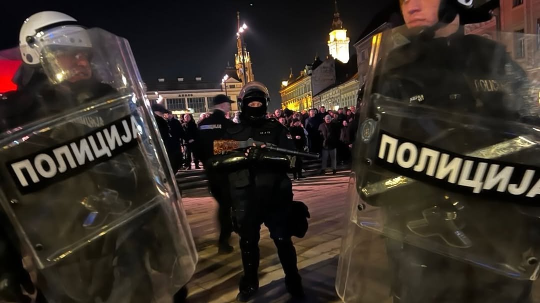 Riot police with shields marked 'ПОЛИЦИЈА' at night during protest