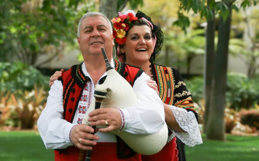 Болгарский фольклор зазвучит в Сан-Диего: концерт четы Варимезовых Иван и Цветанка Варимезови в традиционных болгарских костюмах с гайдой на фоне зелёного парка