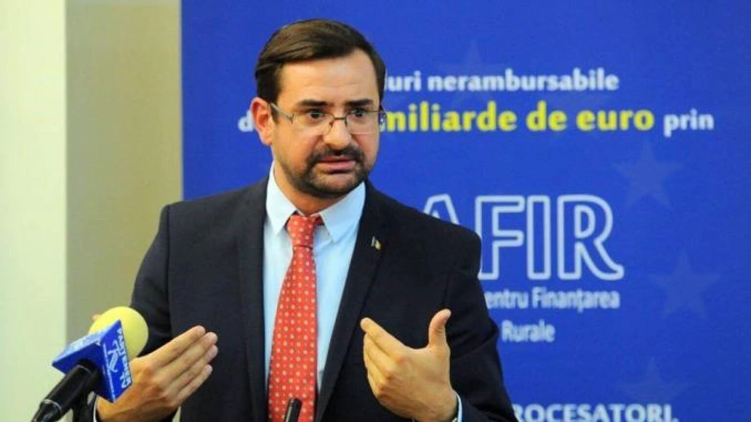 Адриан Кесною выступает на мероприятии AFIR (Агентство по финансированию инвестиций в сельской местности), жестикулирует перед аудиторией с микрофоном, в тёмном костюме с красным галстуком. Фото: AFIR