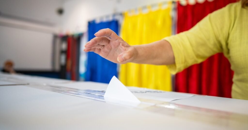Избиратель опускает бюллетень в урну на избирательном участке на фоне занавесок в цветах румынского триколора