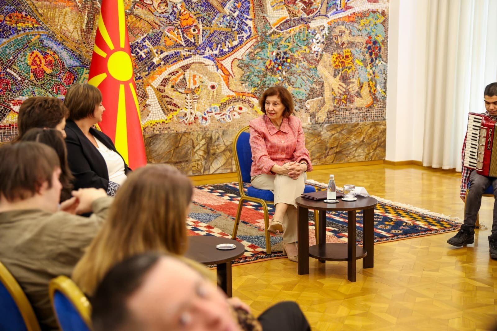 Президент в розовом жакете сидит и слушает молодого человека, играющего на аккордеоне в зале президентского дворца