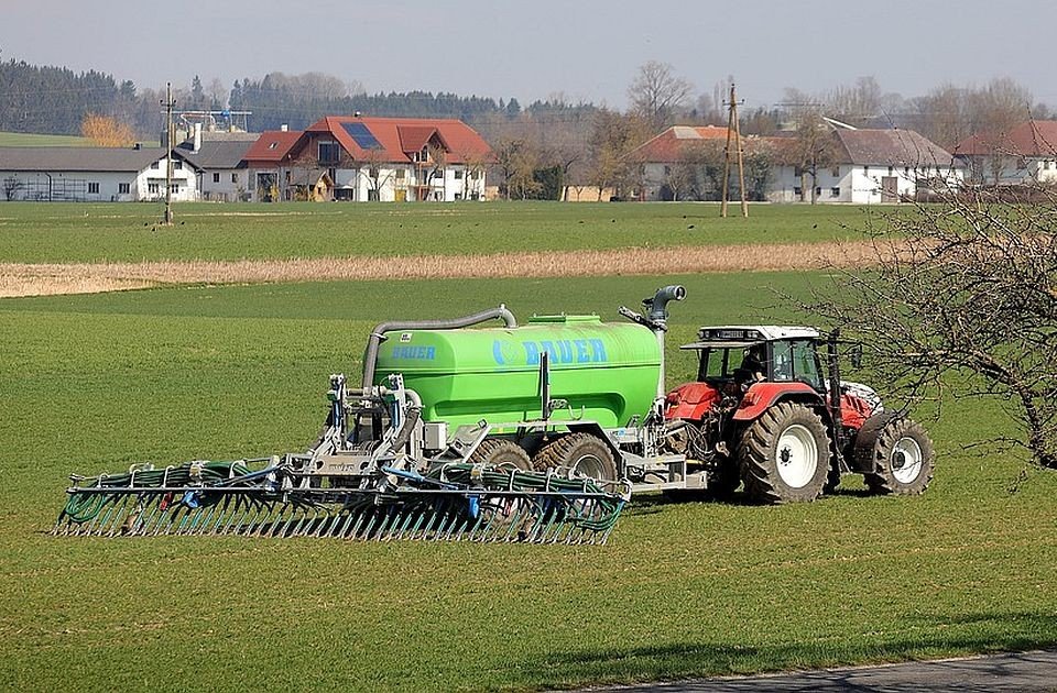Трактор с цистерной для внесения удобрений работает на зелёном поле на фоне сельского пейзажа