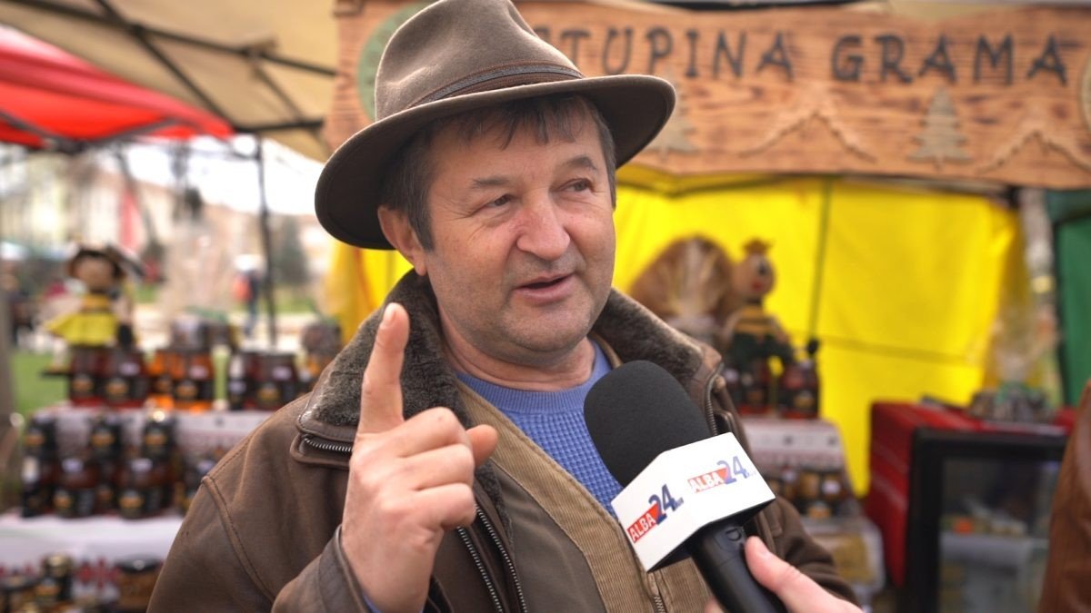 Мужчина в шляпе и кожаной куртке даёт интервью перед стендом с банками мёда и вывеской «Ступина Грама»
