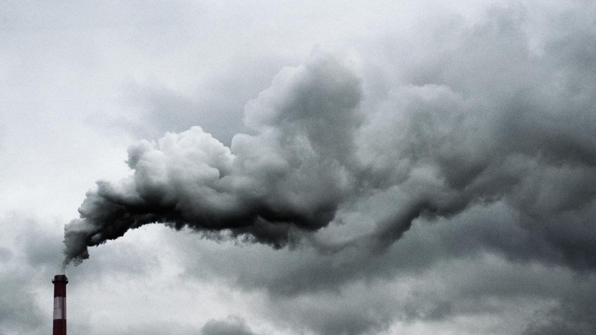 Промышленная дымовая труба выбрасывает густой тёмный дым на фоне пасмурного неба