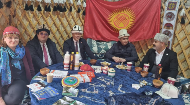 Гости внутри традиционной кыргызской юрты за столом с национальными блюдами, сувенирами и флагом Кыргызстана на выставке в Баку