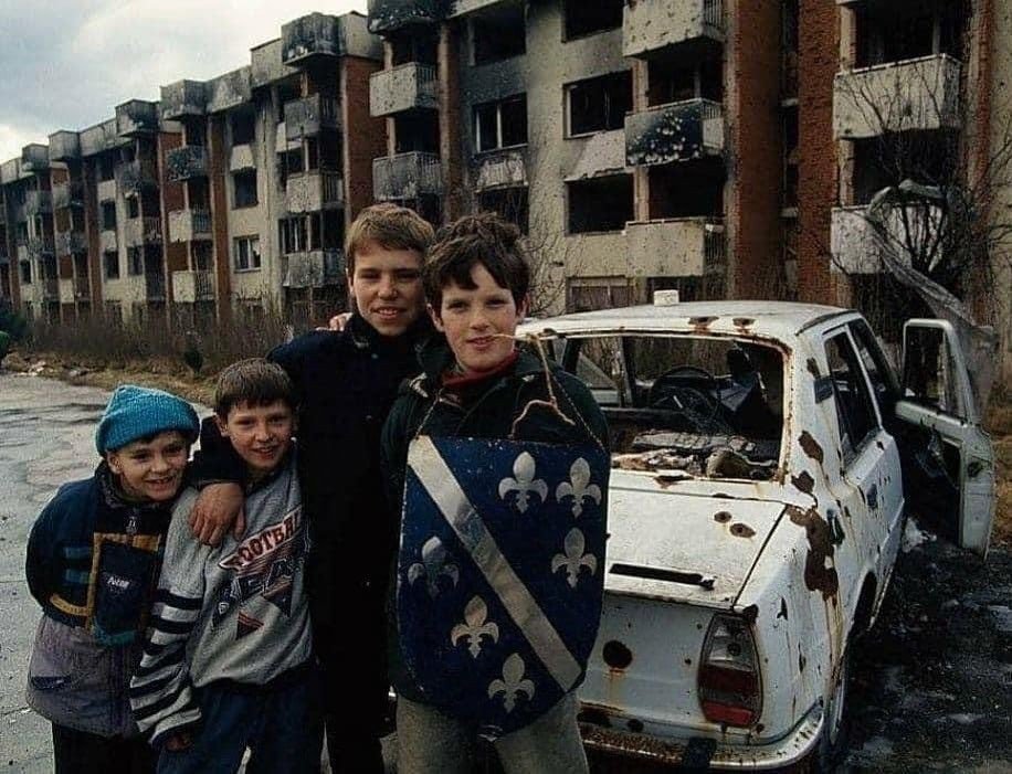 Дети осаждённого Сараево на фоне разрушенных многоэтажек и сгоревшего автомобиля. Один из мальчиков держит щит с гербом Боснии и Герцеговины